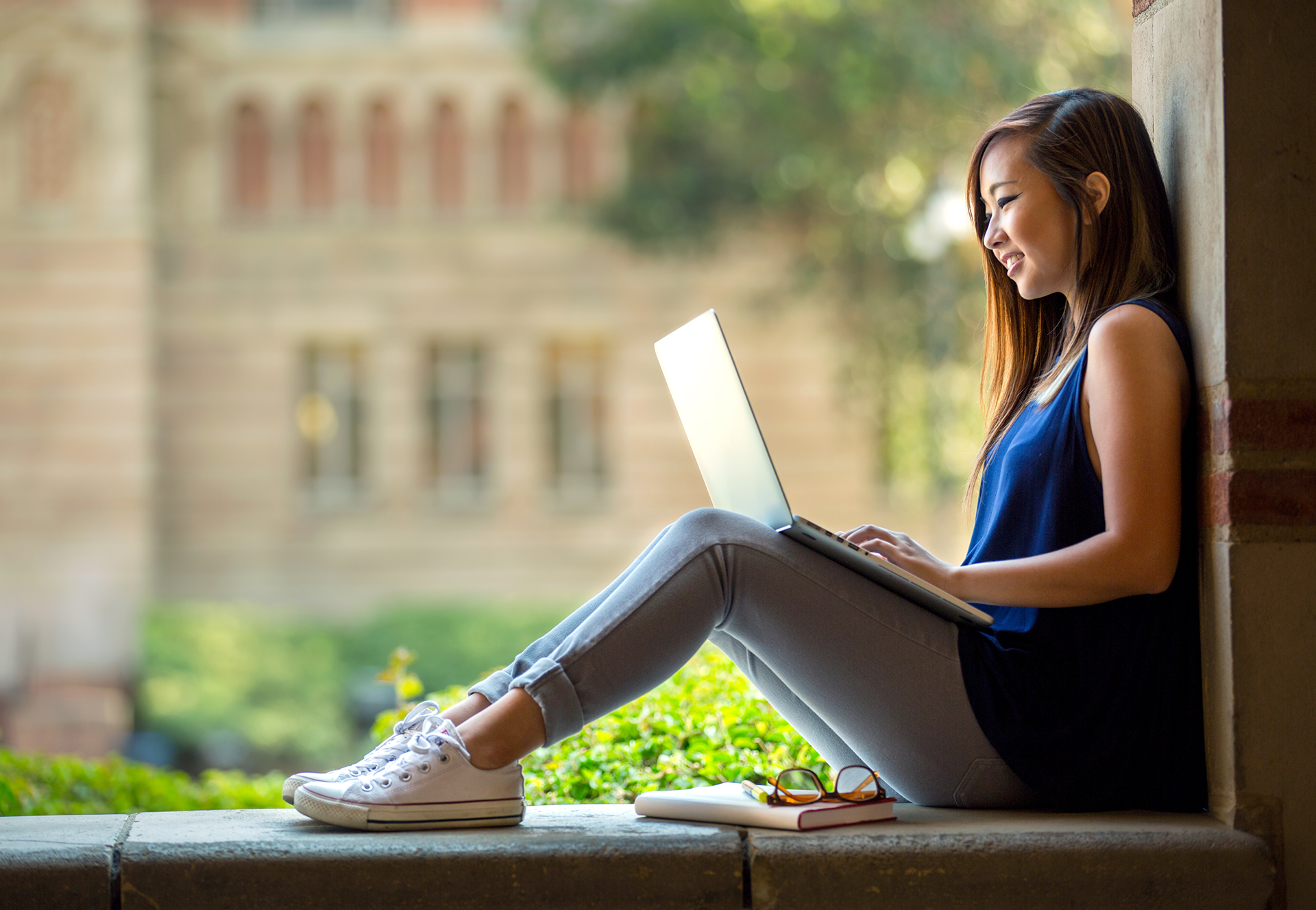 Smiling young Asian woman using laptop outside a college university.