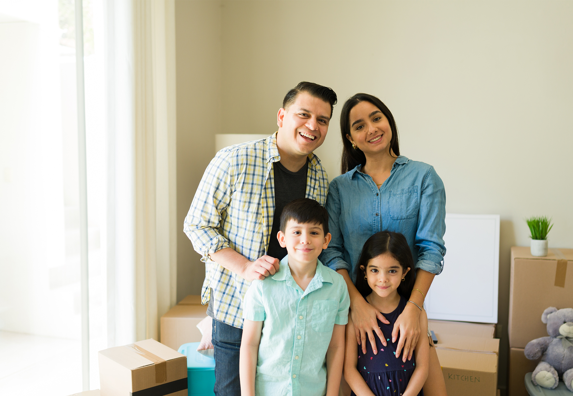 Happy Hispanic couple with two kids smiling in new home with moving boxes on floor in background.