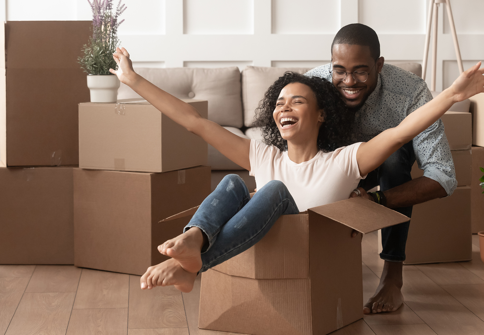 Happy, young African American couple first time homeowners unpacking laughing on moving day. Excited wife riding sitting in cardboard box while husband pushes it.