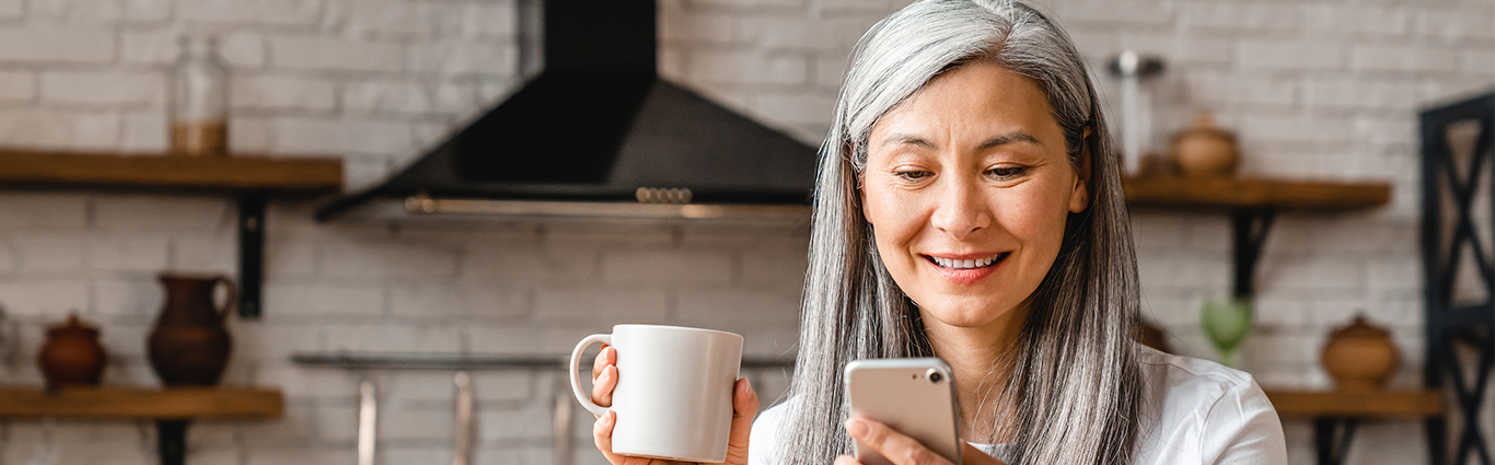 Smiling middle-aged Asian woman drinking coffee in kitchen while looking at smartphone