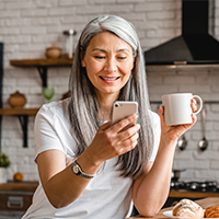 Smiling middle-aged Asian woman drinking coffee in kitchen while looking at smartphone